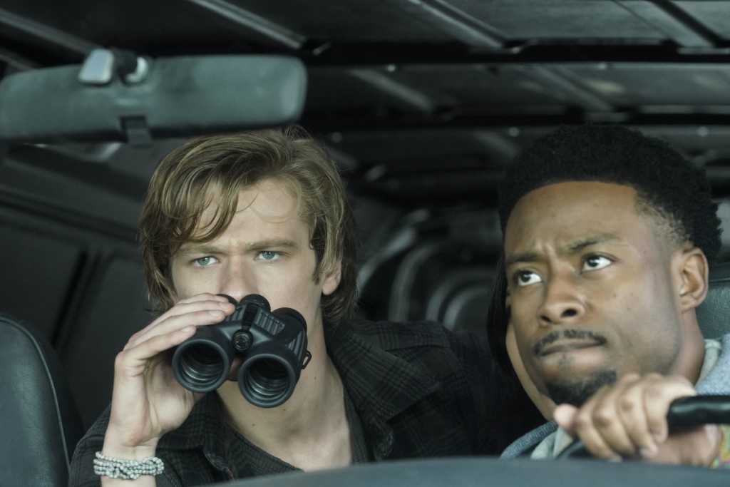 Bozer (Justin Hires) est au volant d'une voiture et Mac (Lucas Till) utilise une paire de jumelles pour surveiller un ennemi.