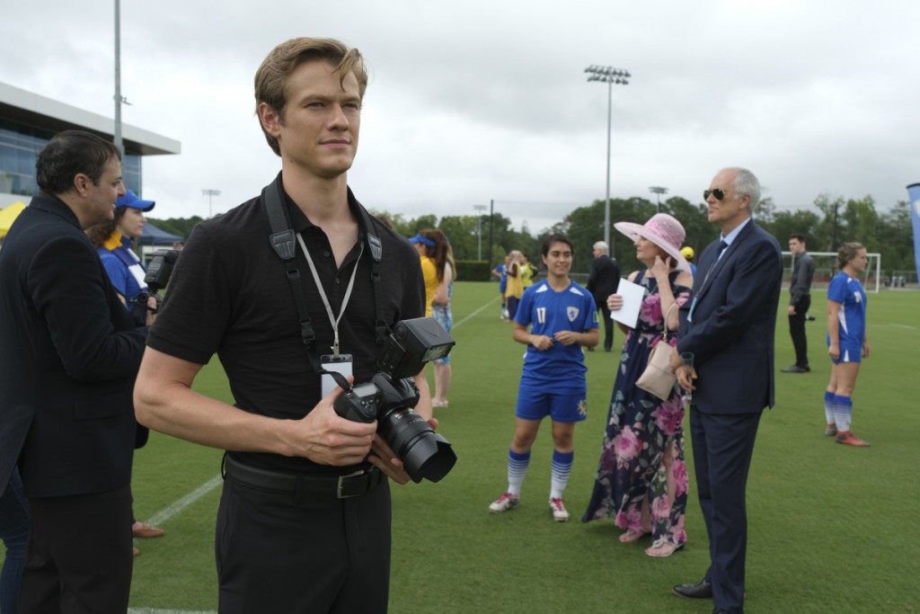 Mac (Lucas Till) se trouve sur un terrain de football avec un appareil photo.